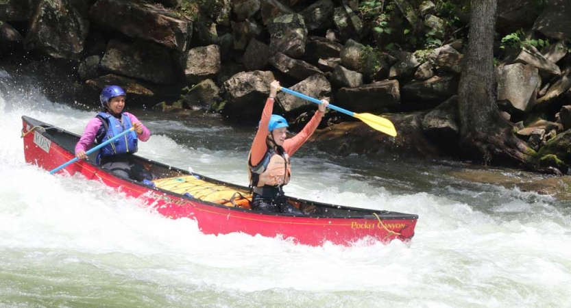 Two people wearing safety gear paddle a canoe through whitewater. The person in the front of the canoe raises their paddle into the air in apparent celebration. 